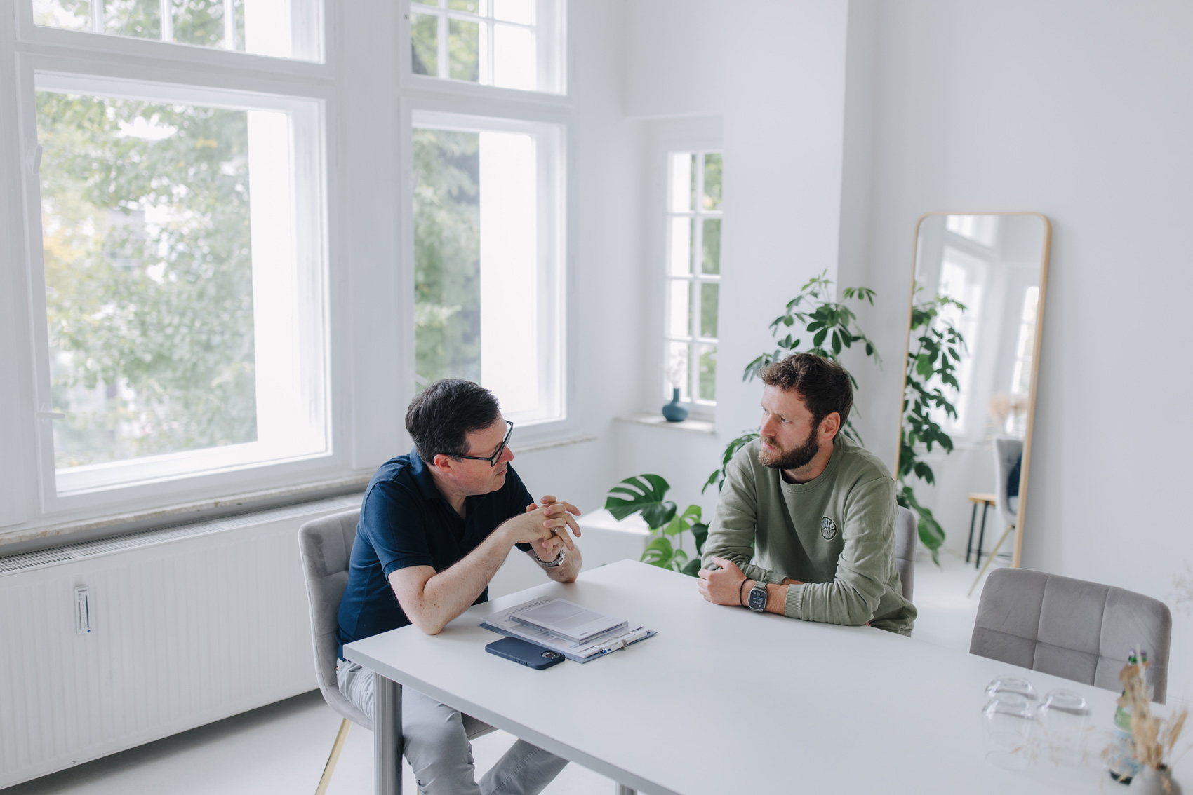 Zwei Männer in einem modernen, hellen Büro bei einem Gespräch am Tisch, Fokus auf Kommunikation und professionellen Austausch.