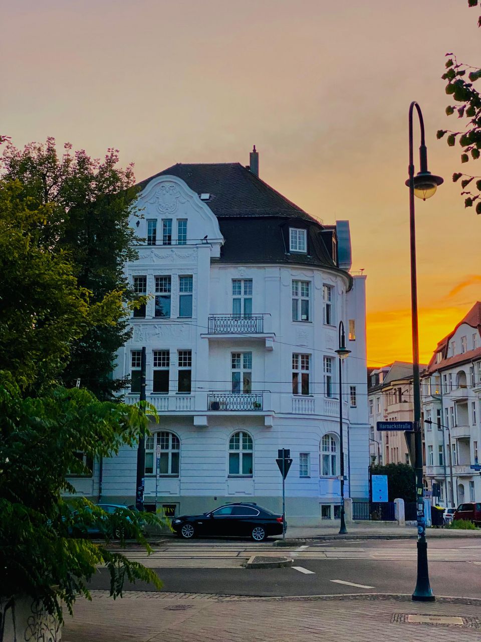 Historisches weißes Gebäude in urbaner Umgebung bei Sonnenuntergang, mit Straßenlaternen und Bäumen im Vordergrund.