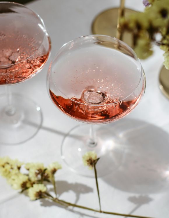 Zwei Gläser mit roséfarbenem Sekt auf weißer Oberfläche, dekoriert mit Blumen und goldenen Details, elegant und festlich.