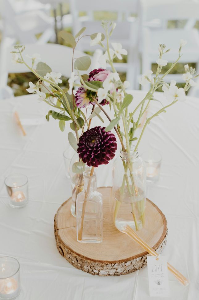 Rustikale Tischdekoration mit Blumen in Glasvasen auf Holzscheibe, umgeben von Teelichtern, für eine elegante Feier.