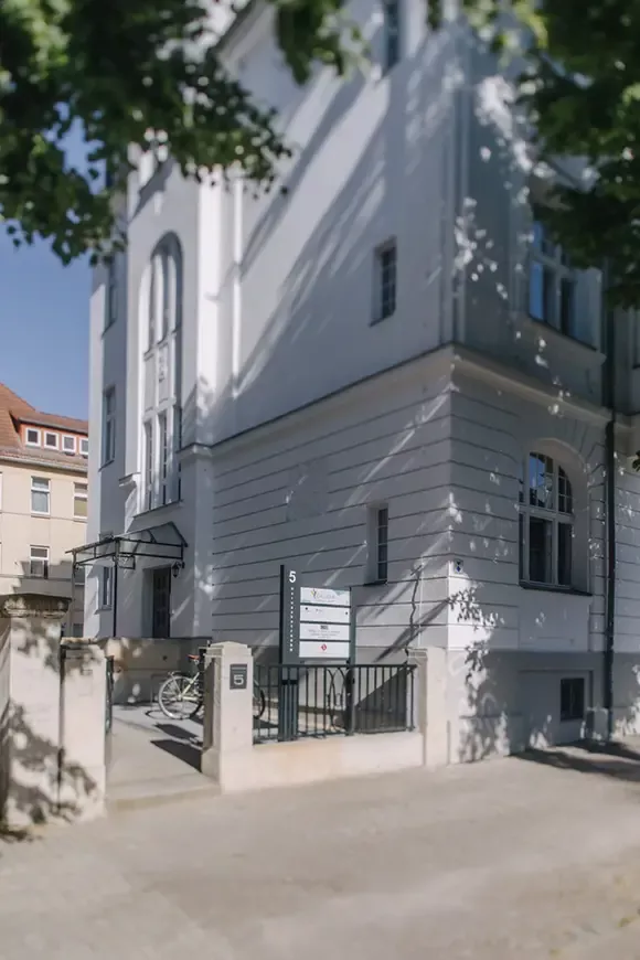 Außenansicht eines historischen Gebäudes mit weißer Fassade, Schildern am Eingang und schattenspendenden Bäumen.