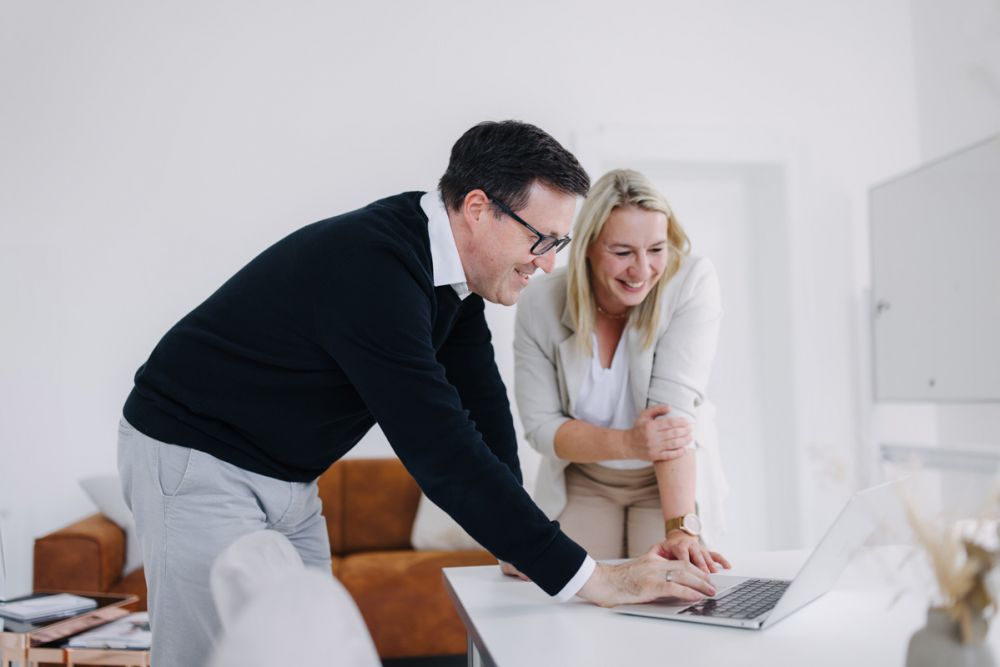 Zwei Kollegen in einem modernen Büro arbeiten gemeinsam an einem Laptop, lächeln und diskutieren konzentriert ein Projekt.