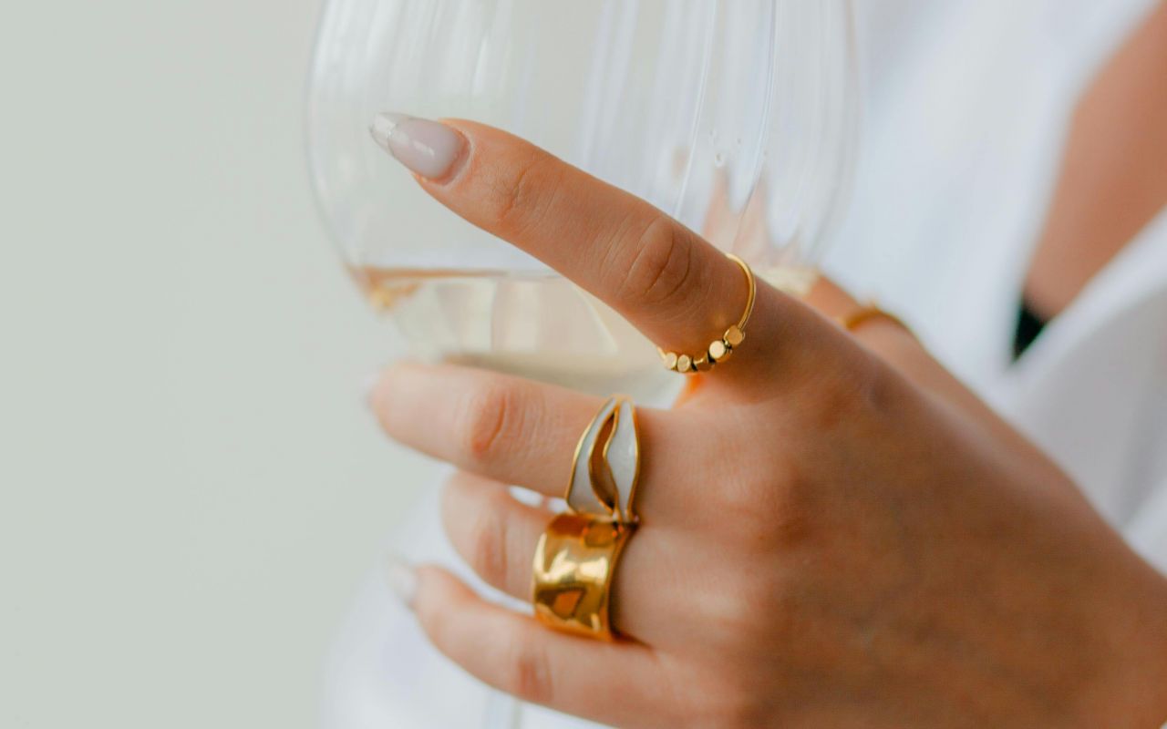 Hand mit elegantem Goldschmuck hält ein Weinglas, stilvoll und minimalistisch inszeniert für einen luxuriösen Lifestyle.